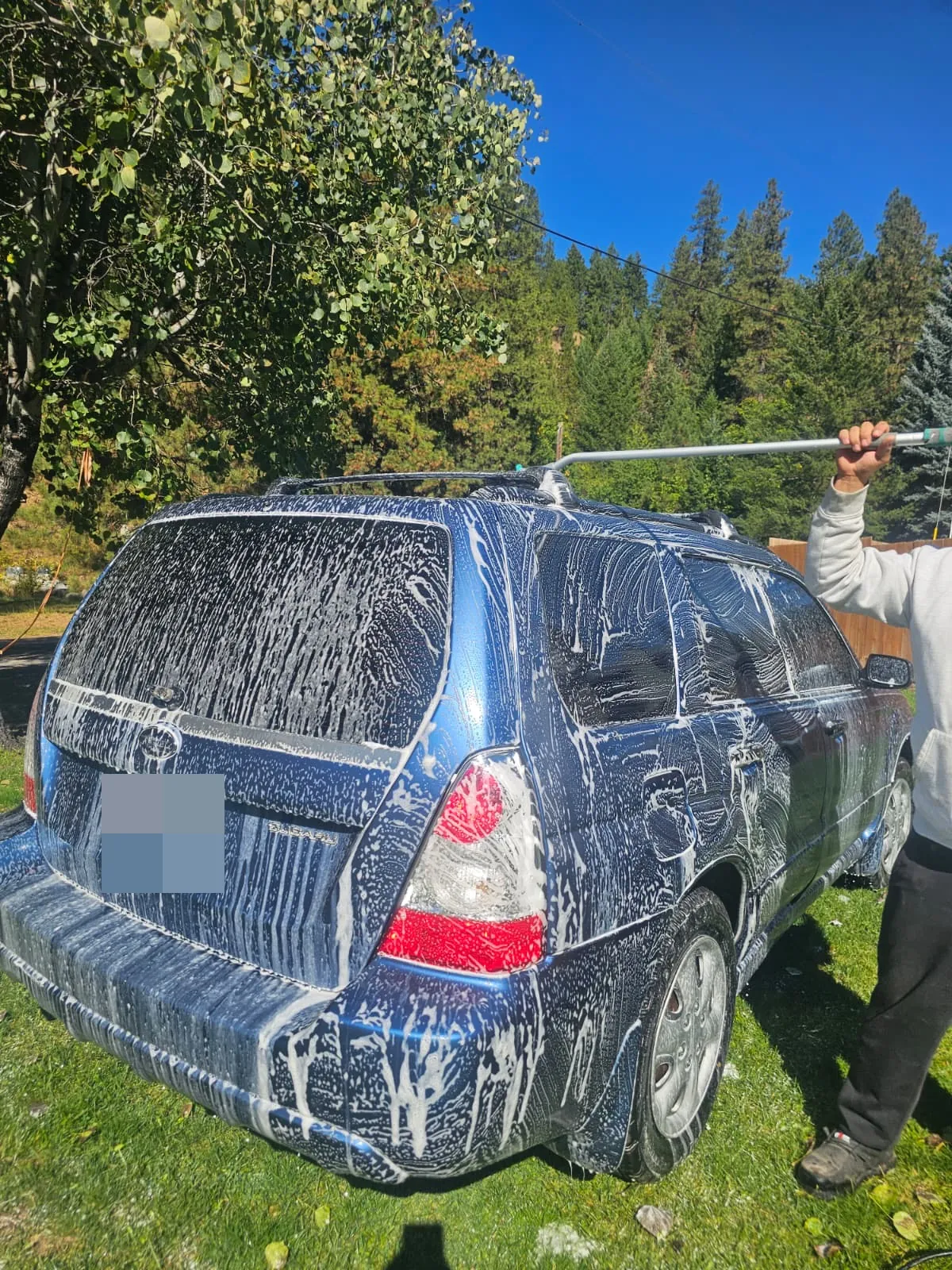 Blue Subaru Forester covered in wash foam, with a person using a long-handled brush